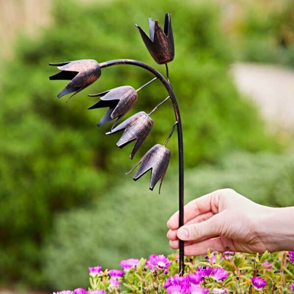 A hand holds a Recycled Metal Echinacea Silhouette Garden Stake shaped like five tulip-like flowers on a stem, with purple blooms and greenery in the background.