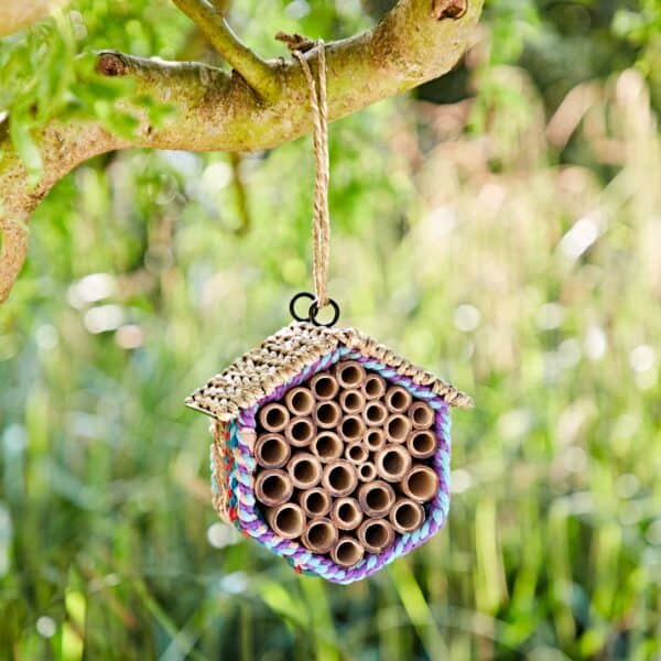 The Hexagonal Recycled Cotton Insect & Bee House, featuring bamboo tubes and straw, hangs from a tree branch with twine against a blurred green outdoor background.