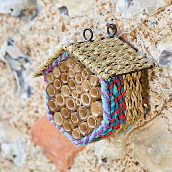 The Hexagonal Recycled Cotton Insect & Bee House, crafted with bamboo tubes and woven straw, is mounted on a rough stone wall.