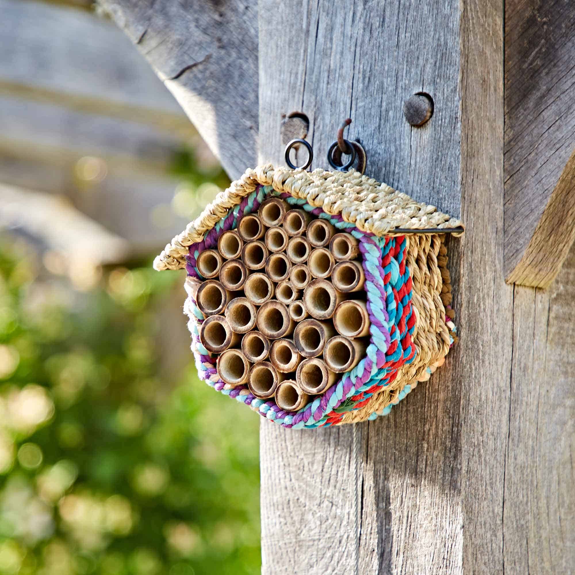 The Hexagonal Recycled Cotton Insect & Bee House features bamboo tubes and woven material, hanging outdoors on a wooden post.
