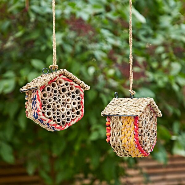 Two small Hexagonal Recycled Cotton Insect & Bee Houses hang from ropes outdoors, featuring bamboo tubes and set against a backdrop of green leaves.