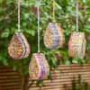 Four Teardrop Recycled Cotton Insect & Bee Houses with bamboo tubes and colourful woven sides hang outside, set against lush green foliage and a wooden fence.