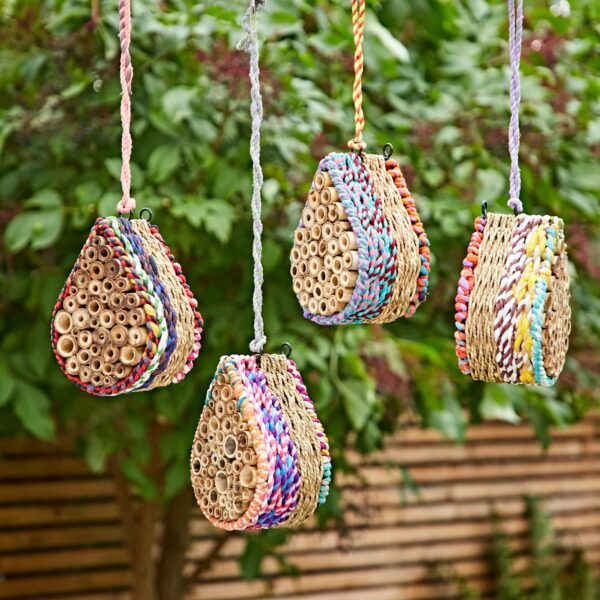 Four Teardrop Recycled Cotton Insect & Bee Houses with bamboo tubes and colourful woven sides hang outside, set against lush green foliage and a wooden fence.