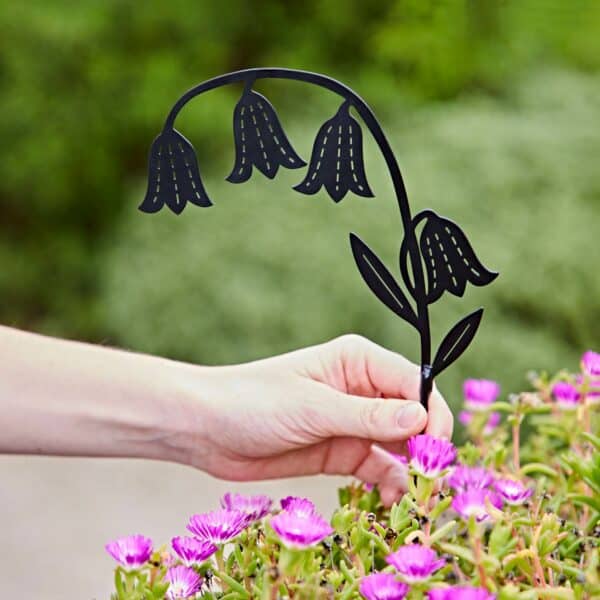 A hand holds a Recycled Metal Echinacea Silhouette Garden Stake, shaped like a bellflower, above blooming pink flowers and lush green foliage.