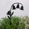 The Recycled Metal Bluebell Silhouette Garden Stake, featuring four drooping blooms, is displayed among green and light grey plants in front of a plain white wall.