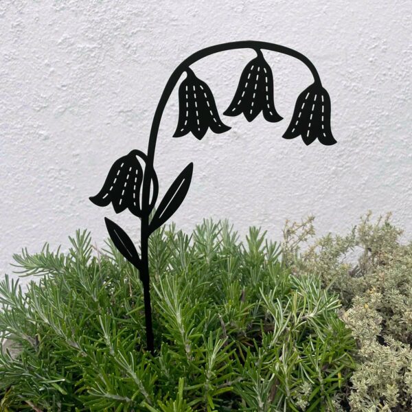 The Recycled Metal Bluebell Silhouette Garden Stake, featuring four drooping blooms, is displayed among green and light grey plants in front of a plain white wall.