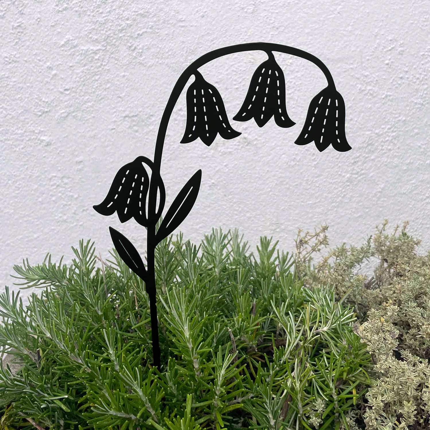 The Recycled Metal Bluebell Silhouette Garden Stake, featuring four drooping blooms, is displayed among green and light grey plants in front of a plain white wall.