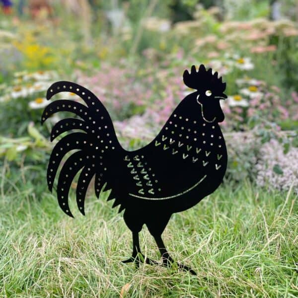 The Recycled Metal Cockerel Silhouette Garden Stake features decorative cutouts and stands on grass among garden flowers, crafted from recycled metal.
