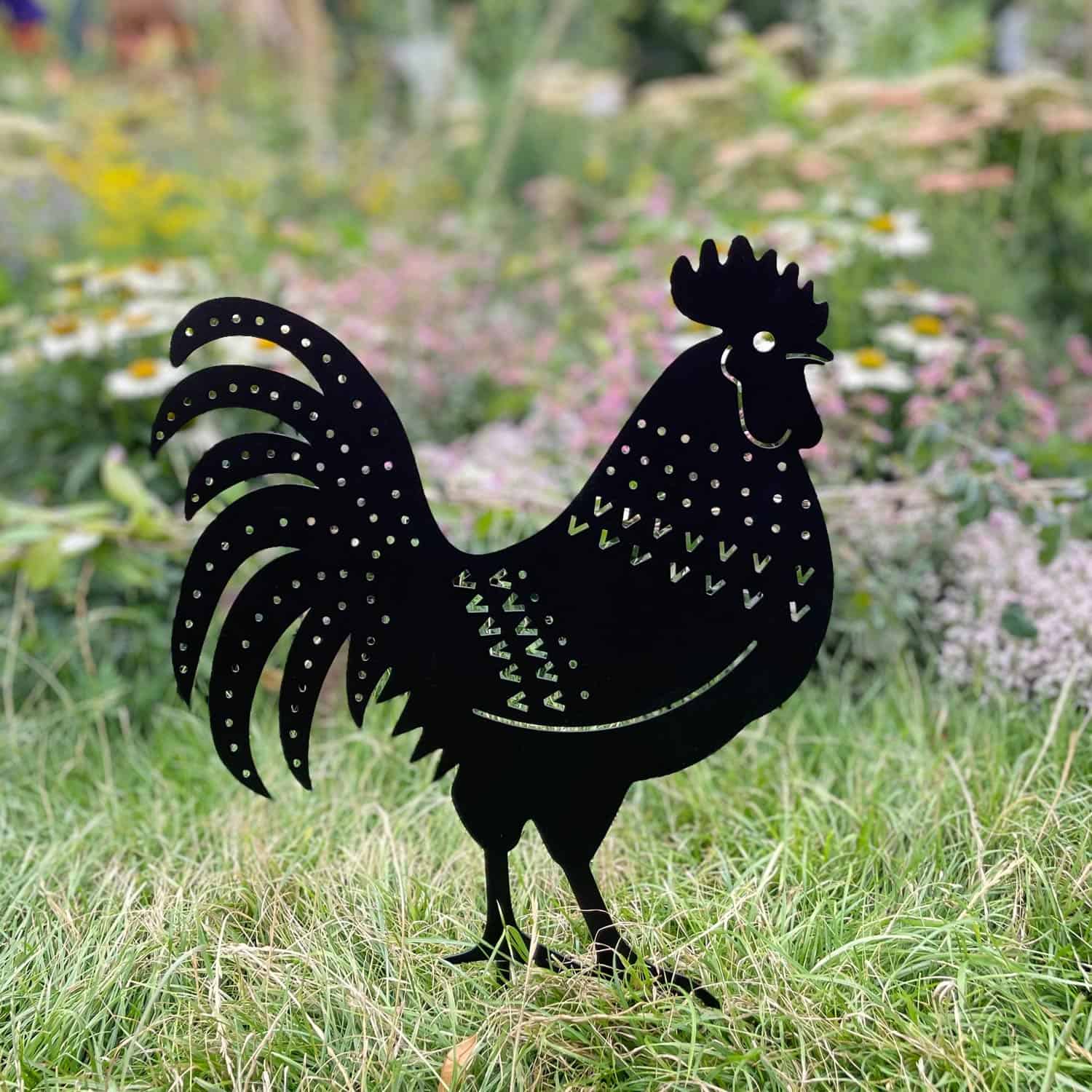 The Recycled Metal Cockerel Silhouette Garden Stake features decorative cutouts and stands on grass among garden flowers, crafted from recycled metal.