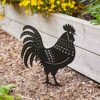 The Recycled Metal Cockerel Silhouette Garden Stake is placed in mulch beside a wooden raised flower bed with yellow flowers.