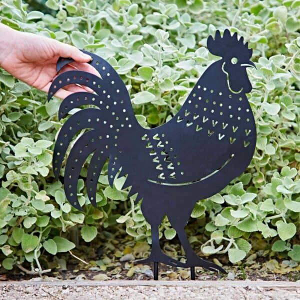 A hand holds a Recycled Metal Echinacea Silhouette Garden Stake with cut-out patterns, displayed against leafy green plants.