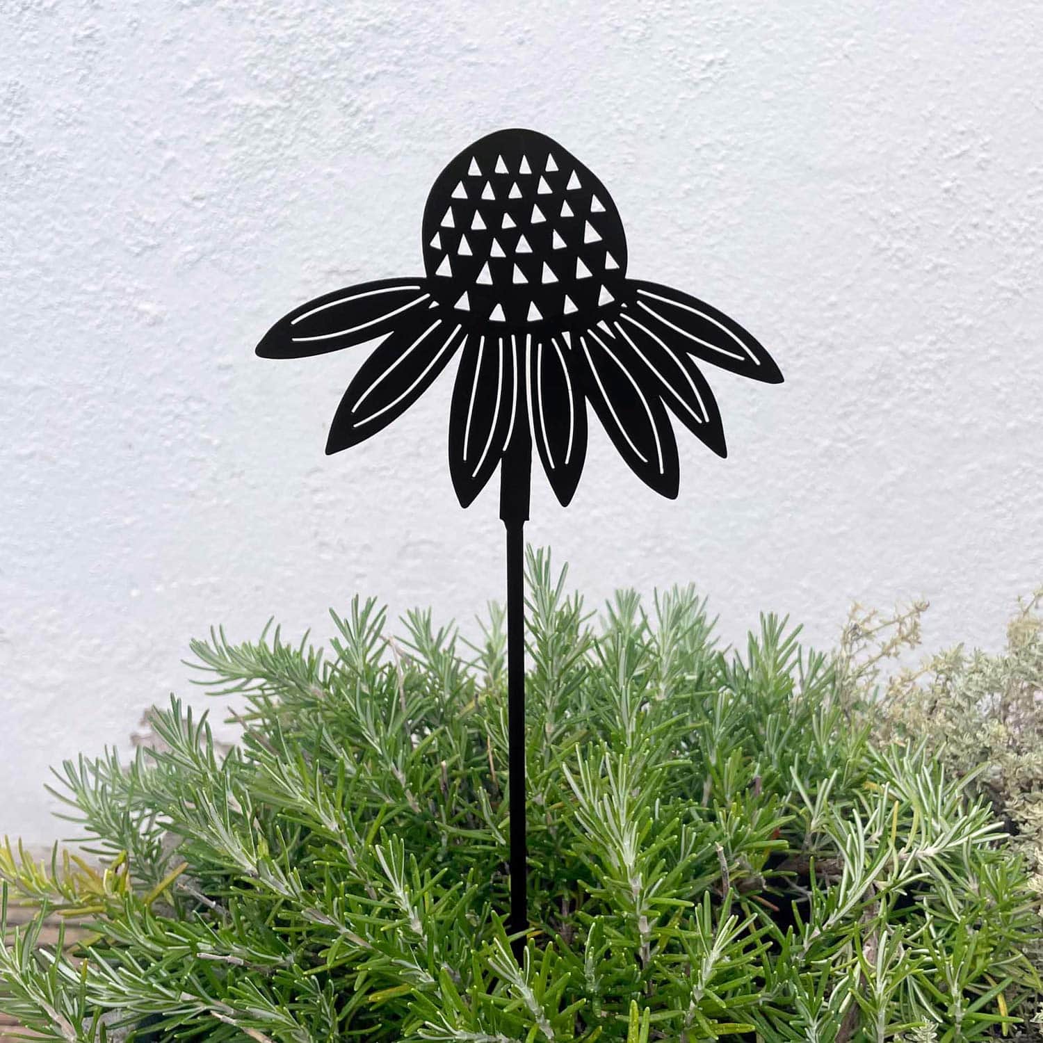 The Recycled Metal Echinacea Silhouette Garden Stake, crafted in black, is set among green foliage with a light-textured wall backdrop.