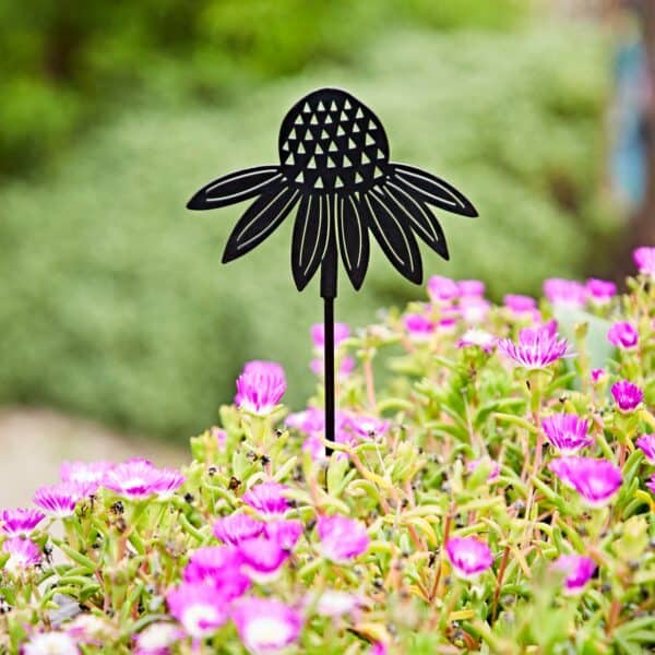 A Recycled Metal Echinacea Silhouette Garden Stake, crafted to resemble a black metal flower, stands gracefully among blooming pink flowers and lush green foliage.