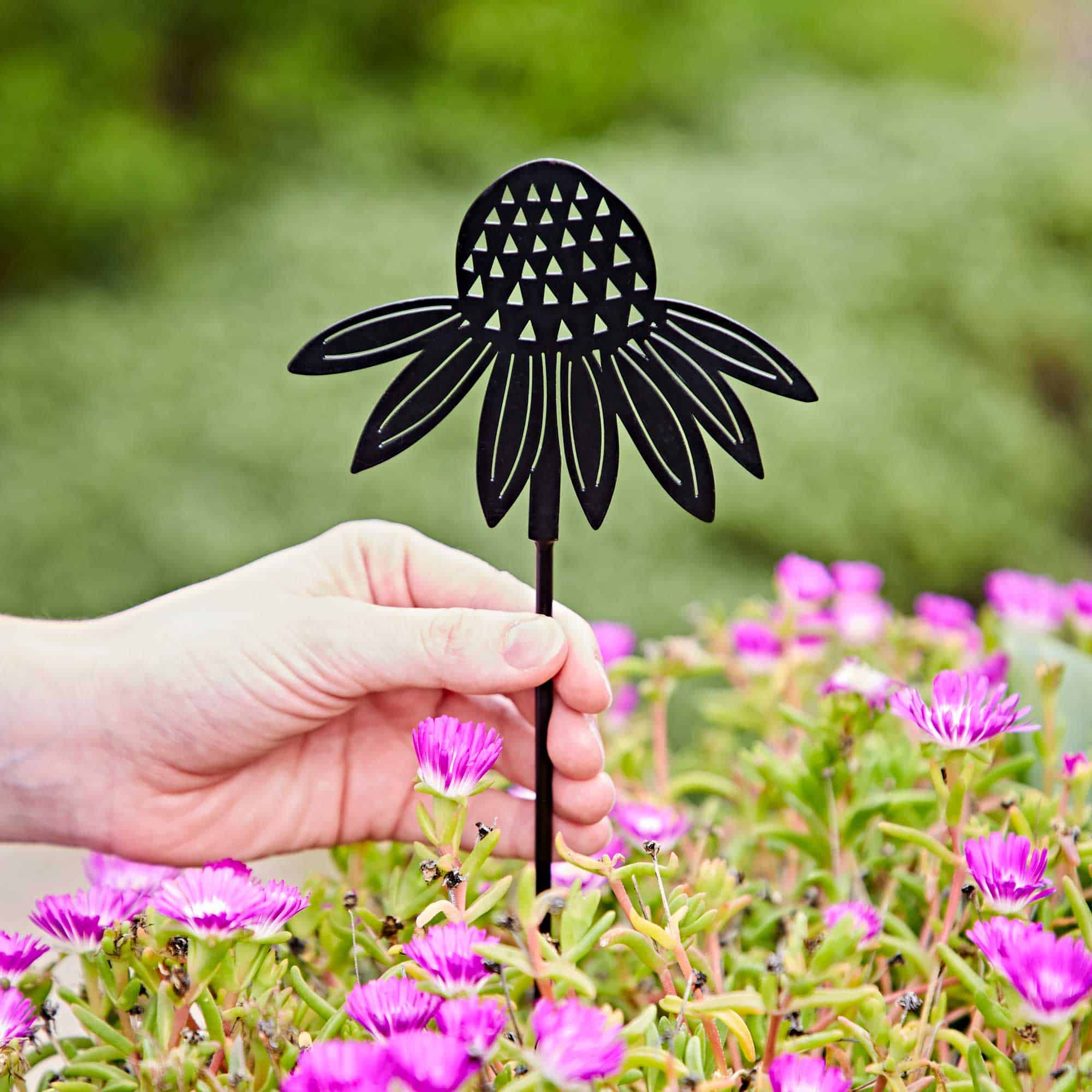 A hand holds a Recycled Metal Echinacea Silhouette Garden Stake shaped like a flower among blooming pink flowers, with green foliage in the background.