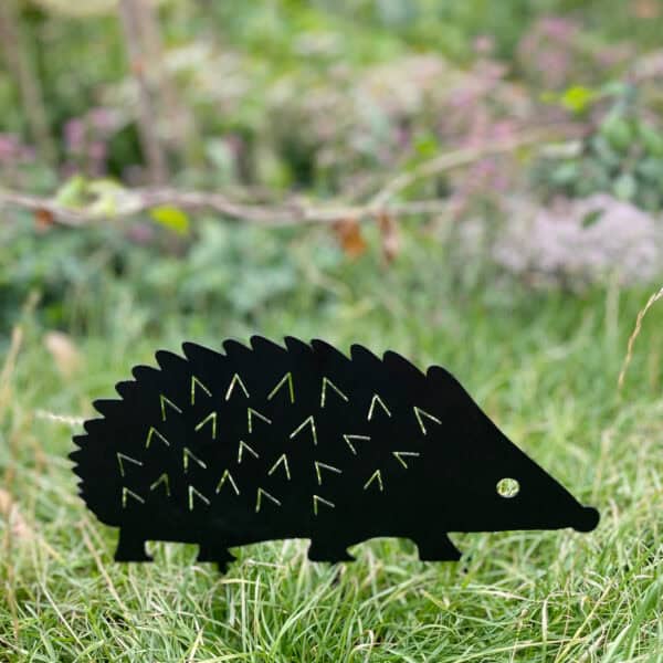 The Recycled Metal Hedgehog Silhouette Garden Stake, crafted from black recycled metal, is shown standing on green grass with a blurred garden background.