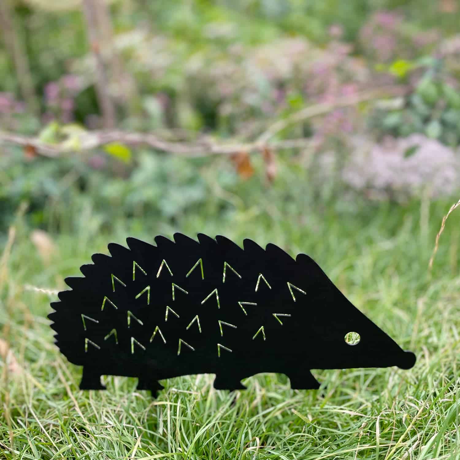 The Recycled Metal Hedgehog Silhouette Garden Stake, crafted from black recycled metal, is shown standing on green grass with a blurred garden background.