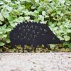 A Recycled Metal Echinacea Silhouette Garden Stake shaped like a hedgehog is displayed in front of green leafy plants on a gravel surface.
