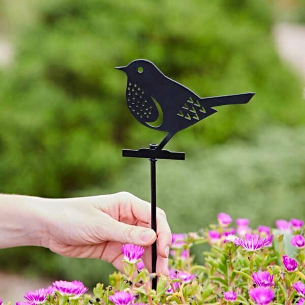 A hand places a Recycled Metal Robin Silhouette Garden Stake among blooming pink flowers outdoors, with lush green foliage in the background.