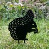 The Recycled Metal Squirrel Silhouette Garden Stake features a black silhouette sitting in grass with trees and pink flowers behind, creating a charming decorative accent for your garden.