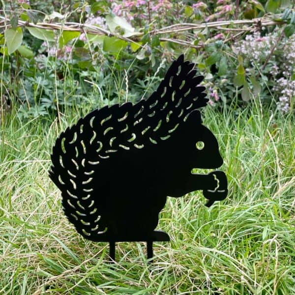 The Recycled Metal Squirrel Silhouette Garden Stake features a black silhouette sitting in grass with trees and pink flowers behind, creating a charming decorative accent for your garden.
