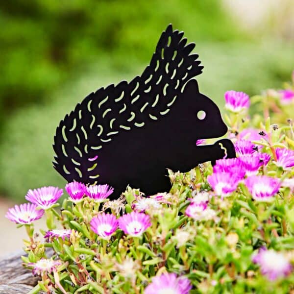 A Recycled Metal Squirrel Silhouette Garden Stake is displayed among blooming pink flowers and vibrant green foliage in an outdoor garden.