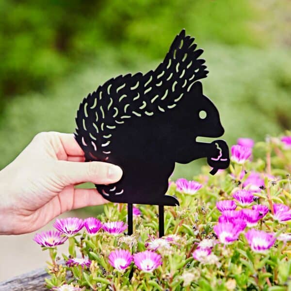 A hand holds a black Recycled Metal Squirrel Silhouette Garden Stake among blooming pink and white flowers, with greenery blurred in the background.