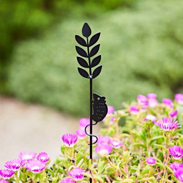 The Recycled Metal Harvest Mouse on Wheat Silhouette Garden Stake, featuring a mouse climbing wheat, decorates your garden beautifully among blooming pink flowers and lush green leaves.