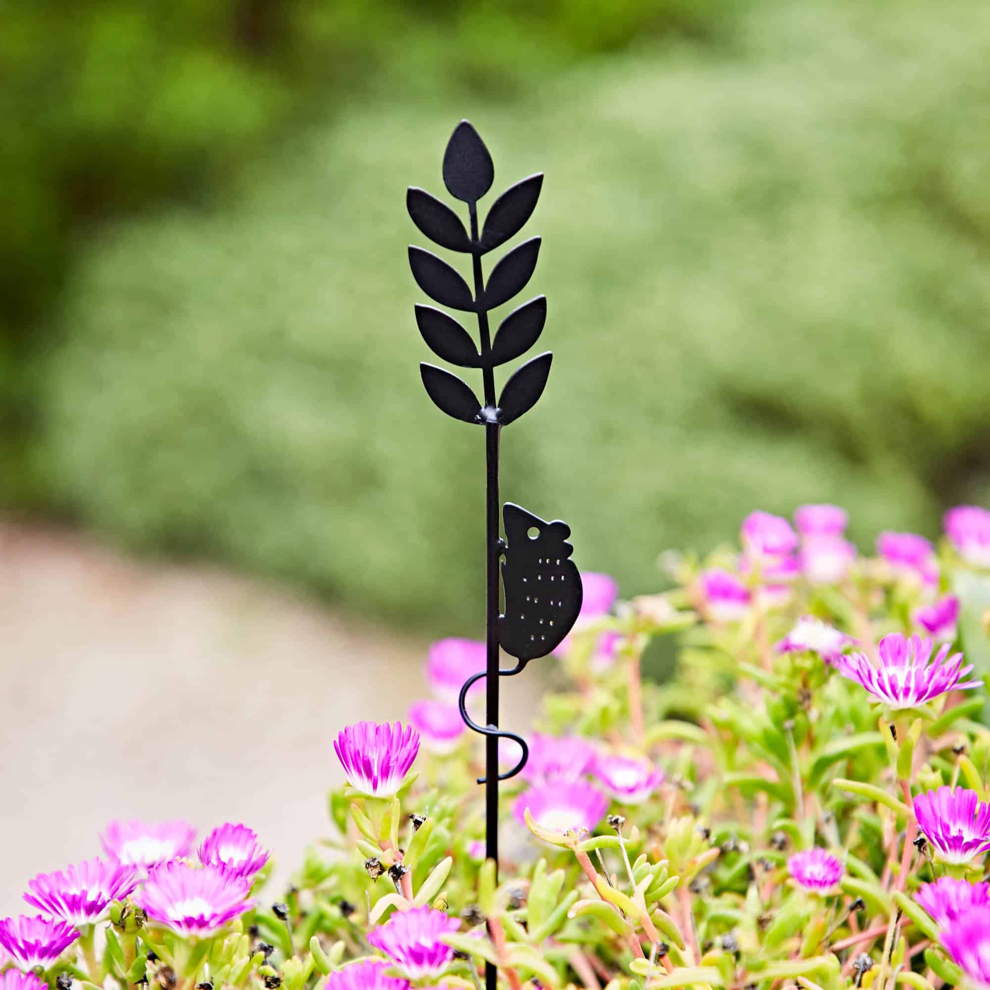The Recycled Metal Harvest Mouse on Wheat Silhouette Garden Stake, featuring a mouse climbing wheat, decorates your garden beautifully among blooming pink flowers and lush green leaves.
