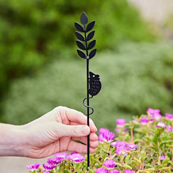 A hand holds the Recycled Metal Harvest Mouse on Wheat Silhouette Garden Stake, featuring a mouse design perched on a plant stem, above blooming pink flowers.