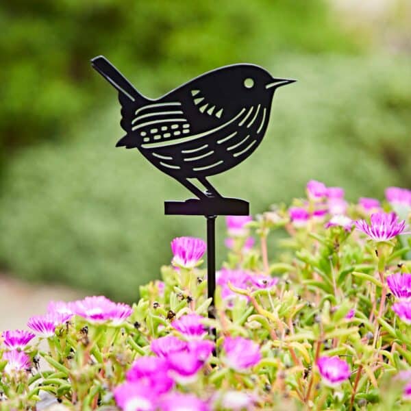 The Recycled Metal Wren Silhouette Garden Stake adds a charming black accent among pink flowers and green foliage, bringing elegance and eco-friendly style to your garden.