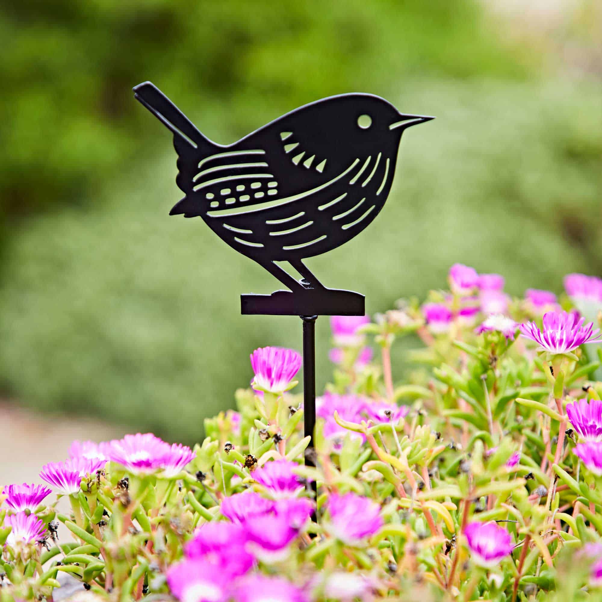 The Recycled Metal Wren Silhouette Garden Stake adds a charming black accent among pink flowers and green foliage, bringing elegance and eco-friendly style to your garden.