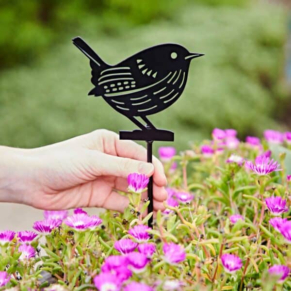 A hand places a black Recycled Metal Wren Silhouette Garden Stake among blooming pink flowers with lush green foliage in the background.