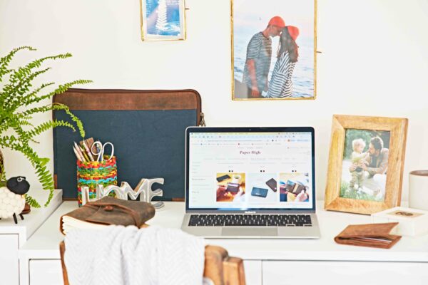 A desk with a laptop, a potted fern reminiscent of the Hampton Court Palace Garden Festival, a pen holder, notebooks, and framed photos of people, set against a white wall.