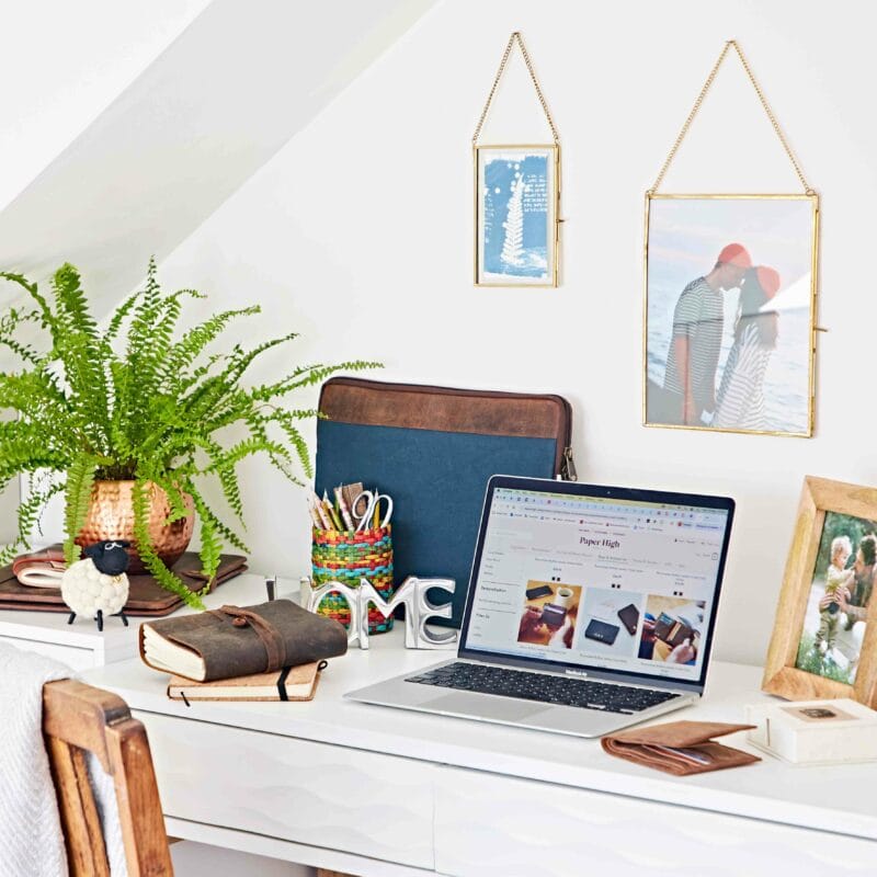 A tidy white desk with a laptop, notebooks, a potted fern reminiscent of the Hampton Court Palace Garden Festival, pens, framed photos, and office supplies under a slanted ceiling.