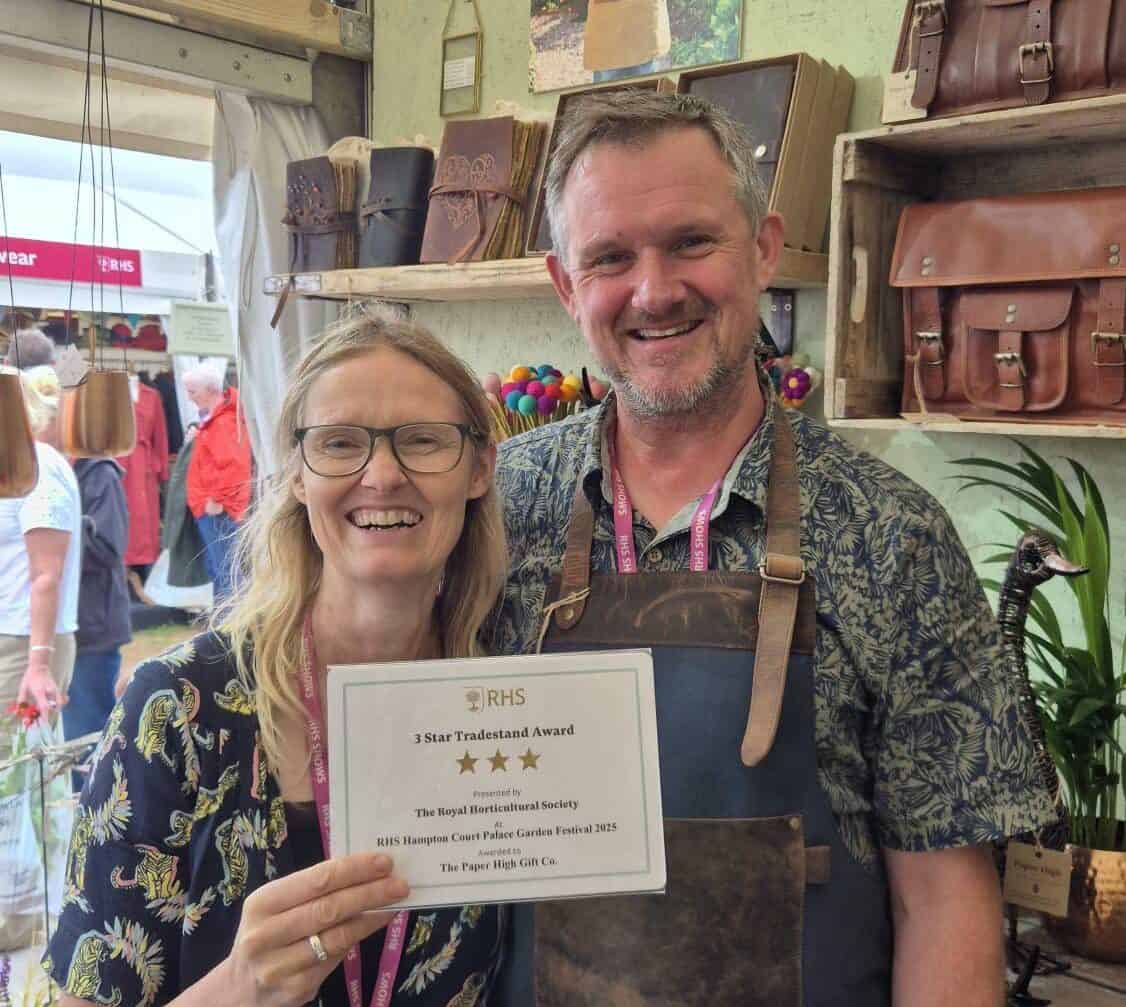 Mark and Claire are smiling at the camera. Claire is holding a three star tradestand award at the RHS Hampton Court Palace Garden Festival 2025. Shelves behind them display leather bags and unique Elephant Dung products among lush plants. Other people are visible in the background.
