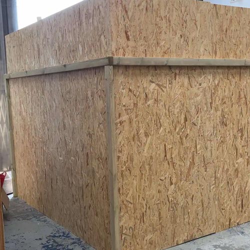 A large, unfinished box structure made of oriented strand board (OSB) panels is positioned indoors on a concrete floor, reminiscent of installations seen at the Hampton Court Palace Garden Festival.