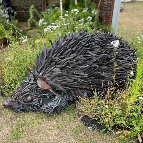 Metal sculpture of a hedgehog made from recycled tools and scrap metal, displayed outdoors among grass and white wildflowers at the Hampton Court Palace Garden Festival.