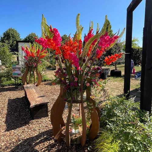A large arrangement of bright pink and orange flowers stands on a wooden display in an outdoor garden area at the Hampton Court Palace Garden Festival on a sunny day.