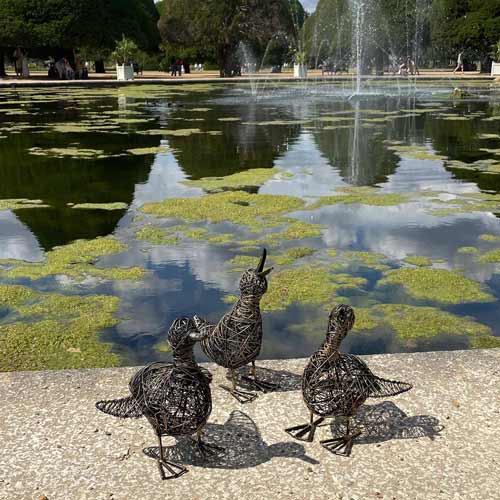 Three wire bird sculptures are positioned on the edge of a pond at the Hampton Court Palace Garden Festival, with green algae, fountains, and trees reflected in the water in the background.
