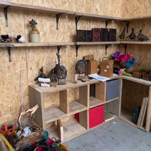 A corner of a workshop or storage area with wooden shelves holding decorative items, books, boxes, and craft supplies against a wood-paneled wall brings to mind the charming displays at the Hampton Court Palace Garden Festival.