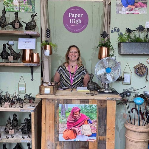 A woman stands smiling behind a wooden counter at a Paper High booth during the Hampton Court Palace Garden Festival, surrounded by metal bird crafts and decorative items.