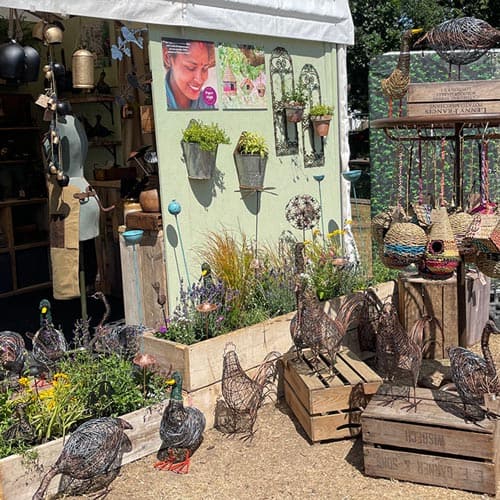 Outdoor garden display inspired by the Hampton Court Palace Garden Festival, featuring wire animal sculptures, potted plants, birdhouses, and hanging decorations in front of a booth with gardening items and a photo of a smiling person on the wall.