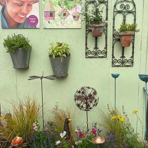 Metal flower pots and hanging plant holders decorate a pale green wall, evoking the charm of the Hampton Court Palace Garden Festival; garden ornaments and colorful flowers bloom at the base.