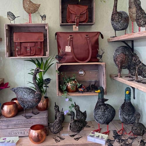 Wire bird sculptures, leather bags, and copper bowls displayed on wooden shelves and crates at the Hampton Court Palace Garden Festival, with potted plants and decorative items arranged around them.
