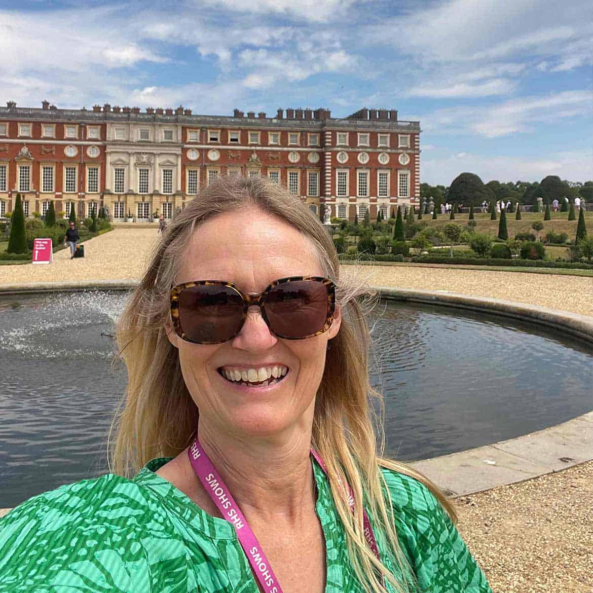 A woman wearing sunglasses and a green top takes a selfie in front of a circular fountain, with the historic Hampton Court Palace and manicured gardens—home to the famous Garden Festival—in the background.