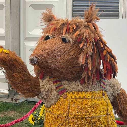 A large, furry animal sculpture made from natural materials stands outdoors at the Hampton Court Palace Garden Festival, wearing a yellow garment and reaching out with one arm.