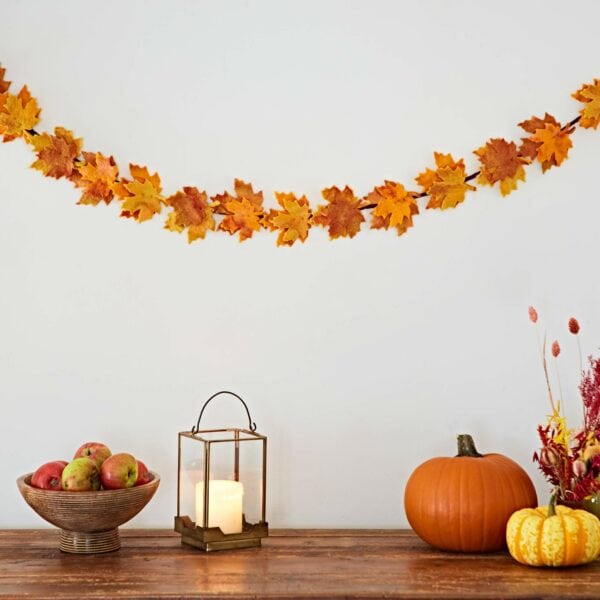 A wooden table features a bowl of apples, a lantern with a lit candle, a pumpkin, small gourds, and a Felt Autumn Leaves Garland hanging on a white wall.