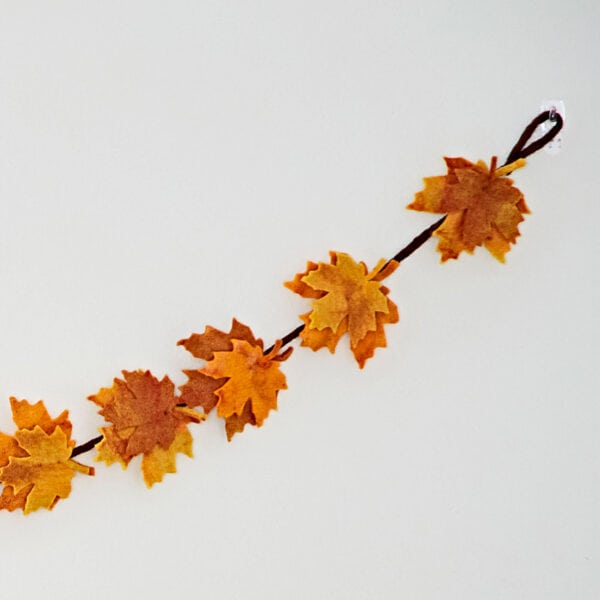 The Felt Autumn Leaves Garland, in orange and yellow shades, hangs on a brown string against a white wall.