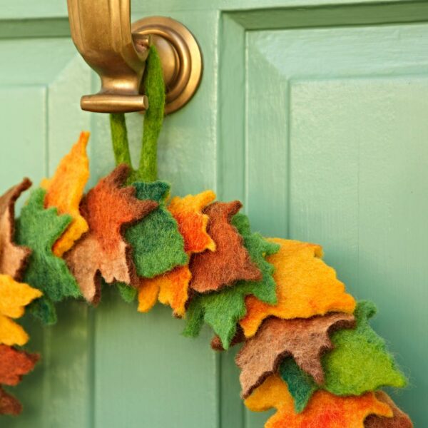 The Felt Woodland Autumn Wreath, made of colourful felt leaf shapes, hangs on a brass door knocker on a light green door, bringing charming autumn decor to your entrance.