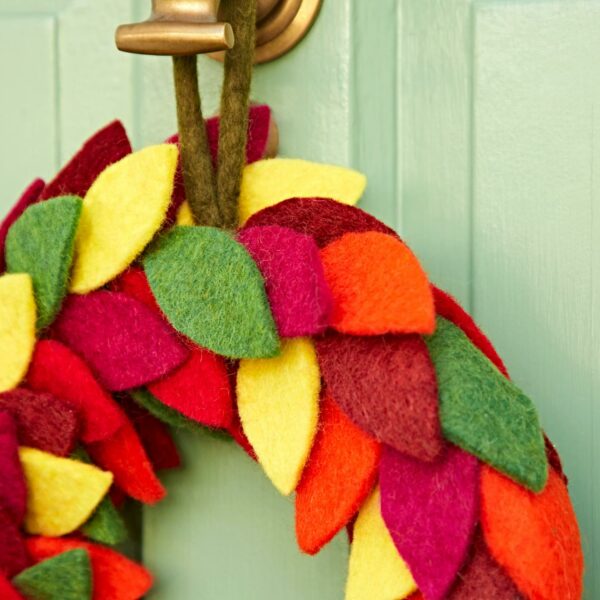 The Felt Colourful Leaf Wreath hangs by a brown strap and brass door knocker on a light green door, displaying its vibrant leaves up close.